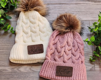 May include: Two knitted winter hats with faux fur pom poms and personalized leather patches. The hats are white and pink with the names "Rylie" and "Kaitlyn" on the patches.