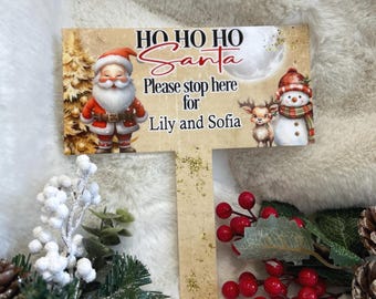 May include: A festive Christmas sign with Santa Claus, a snowman, and a reindeer. The sign says "Ho Ho Ho Santa, Please stop here for Lily and Sofia." The sign is brown with gold glitter and Christmas imagery.