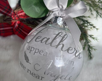 May include: Clear glass Christmas ornament filled with white feathers. The ornament has silver text that reads "Feathers appear when Angels are near." A silver ribbon is tied at the top.