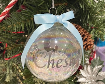 May include: A clear glass Christmas bauble with the word "Chest" in a cursive font. The bauble is filled with iridescent beads and has a light blue ribbon tied in a bow at the top. The bauble hangs on a Christmas tree.