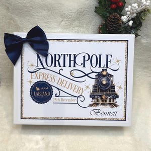May include: A white framed sign with a navy blue bow. The sign reads "NORTH POLE EXPRESS DELIVERY" with a train illustration and "25th December". The name "Bennett" is at the bottom. Gold glitter accents the border and text.