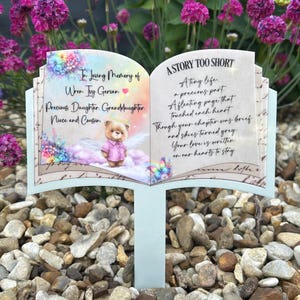 以下が含まれることがあります： 開いた本型の庭の記念碑。左ページには「In Loving Memory of Wren Ty Gervan」と書かれ、右ページには「A Story Too Short」と書かれています。天使の羽を持つテディベアが中央にあり、花柄のアクセントが付いています。