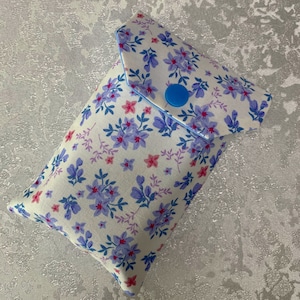 May include: A white fabric pouch with a blue button closure. The pouch is decorated with a floral pattern in shades of blue, pink, and purple.