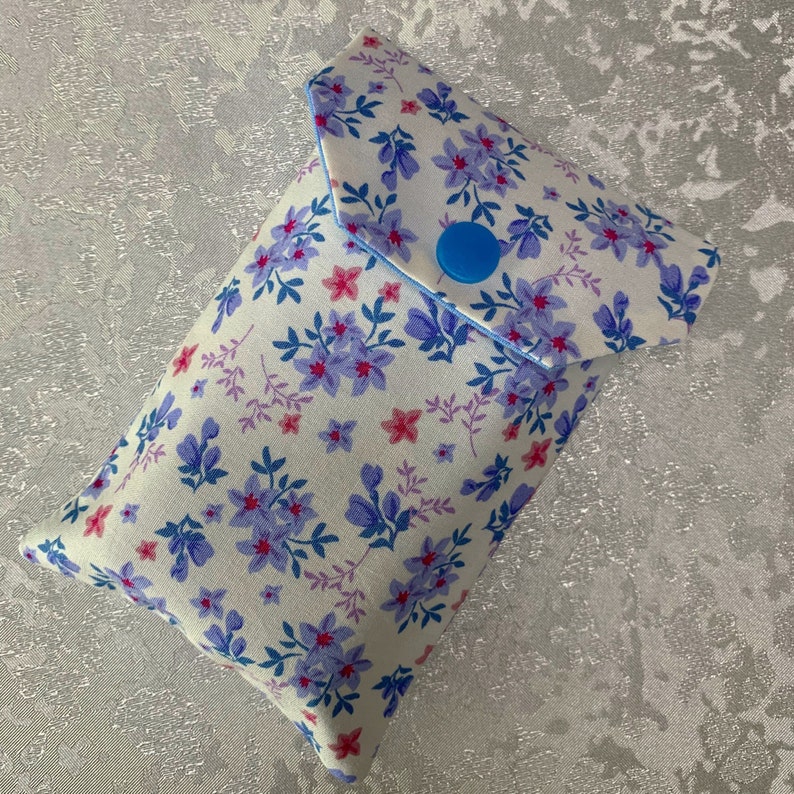 May include: A white fabric pouch with a blue button closure. The pouch is decorated with a floral pattern in shades of blue, pink, and purple.