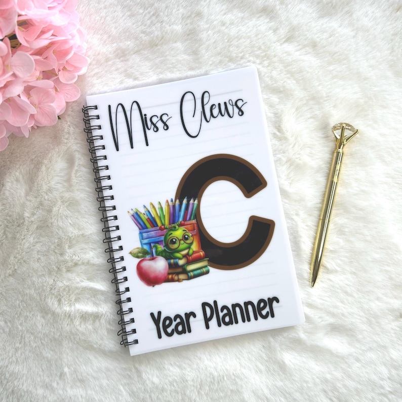 May include: White spiral-bound year planner with the text "Miss Clews" and "Year Planner". The cover features a large brown letter "C", a cartoon frog with glasses, pencils, books, and a red apple. A gold pen is next to the planner.