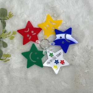 Puede incluir: Un conjunto de llaveros con forma de estrella en rojo, amarillo, verde, azul y blanco. Cada estrella presenta una expresión facial diferente o el texto "HOW DO I FEEL TODAY?". Los llaveros están unidos a un llavero plateado.