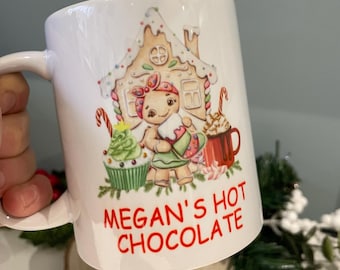 May include: White ceramic mug with a festive design featuring a gingerbread person, a gingerbread house, a cupcake, and a mug of hot chocolate. The mug has the text "Megan's Hot Chocolate" in red.