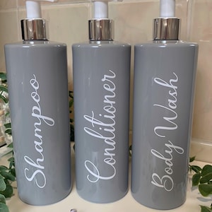 Puede incluir: Tres botellas de bomba grises con etiquetas blancas. Las etiquetas dicen "Champú", "Acondicionador" y "Gel de baño".