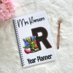 May include: White spiral-bound year planner with the text "Mrs. Ransom" and "Year Planner" in black. The cover features a large brown letter "R" and a colorful illustration of pencils, books, an apple, and a cartoon turtle. A rose gold pen rests beside the planner.