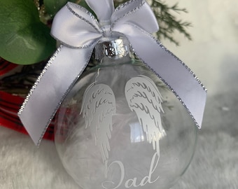 May include: Clear glass Christmas ornament with angel wings and the word "Dad" etched on the front. The ornament is filled with white feathers and has a white ribbon with silver trim. The ornament is tied in a bow.