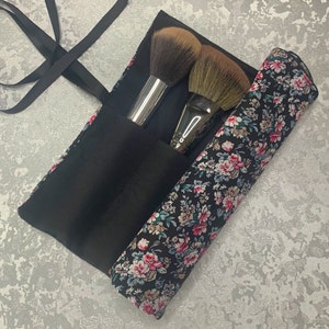 Può includere: Un rotolo nero per pennelli da trucco con stampa floreale. Il rotolo ha due scomparti per pennelli e una chiusura a zip. Il rotolo è aperto e sono visibili due pennelli da trucco.