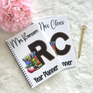 May include: Two white spiral-bound Year Planner notebooks. One notebook has "Mrs Ransom" and a large "R" with a cartoon frog and pencils. The other notebook has "Miss Clews" and a large "C". A gold pen is next to the notebooks.