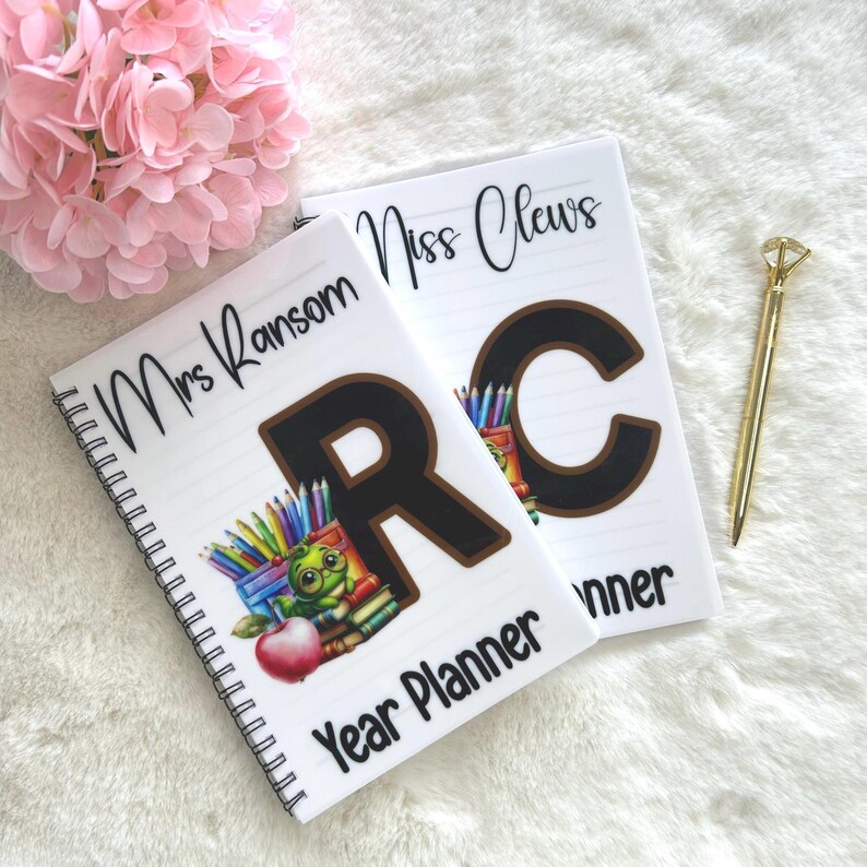 May include: Two white spiral-bound Year Planner notebooks. One notebook has "Mrs Ransom" and a large "R" with a cartoon frog and pencils. The other notebook has "Miss Clews" and a large "C". A gold pen is next to the notebooks.