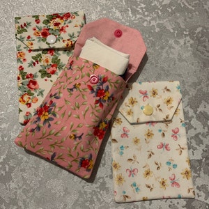 May include: Three fabric pouches with floral patterns. The pouches have a flap closure with a button. The pouch on the left has a pink and white floral pattern, the middle pouch has a pink floral pattern, and the pouch on the right has a white floral pattern.