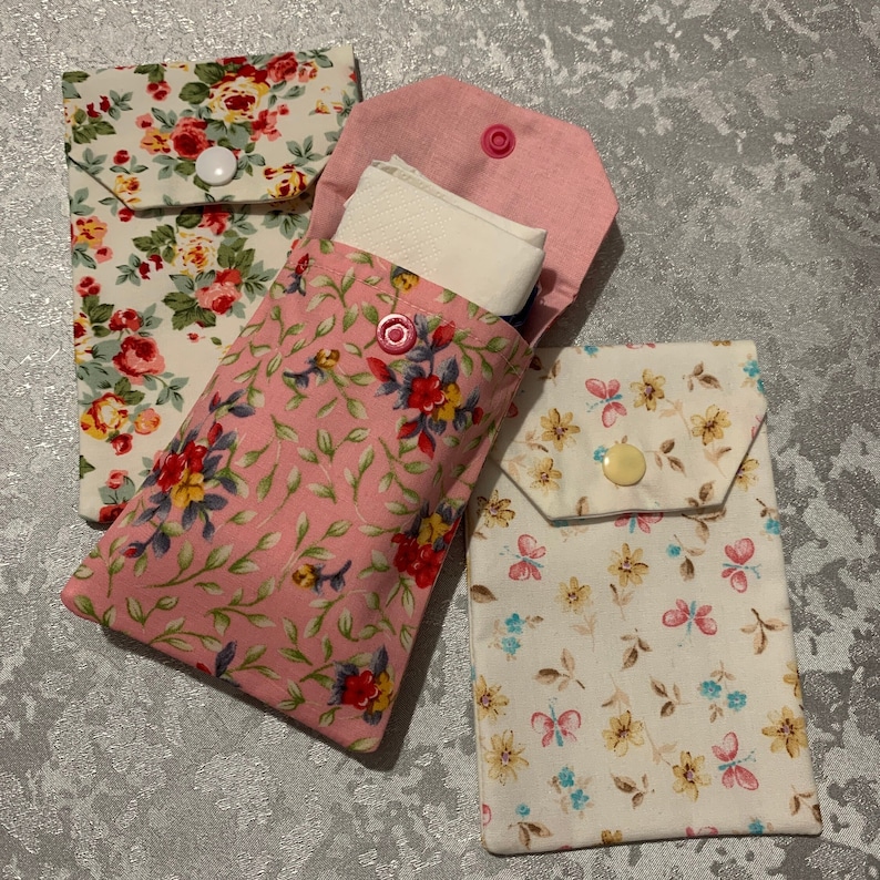 May include: Three fabric pouches with floral patterns. The pouches have a flap closure with a button. The pouch on the left has a pink and white floral pattern, the middle pouch has a pink floral pattern, and the pouch on the right has a white floral pattern.