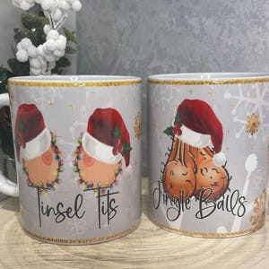 May include: Two white ceramic mugs with festive, humorous designs. One mug displays breasts in a Santa hat with the text "Tinsel Tits". The other mug has testicles in a Santa hat with the text "Jingle Balls". Both mugs have snowflake and gold accents.