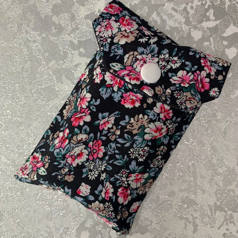 May include: A black fabric pouch with a floral print and a white button closure. The pouch is designed to hold small items like a phone or keys.