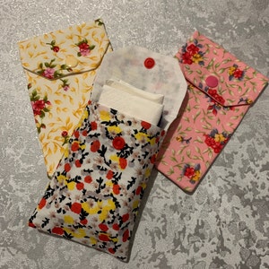 May include: Three fabric pouches with floral patterns. The pouch in the centre is open and has a white tissue inside. The pouches are made of cotton and have a button closure.
