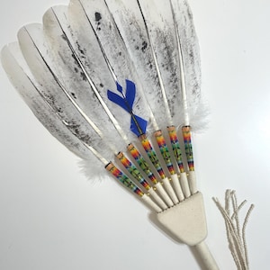 May include: A handcrafted fan featuring white feathers with dark markings. The fan has a cream-colored handle and a decorative blue ribbon. The feather stems are adorned with colorful, patterned accents.