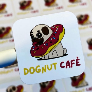 Pu&ograve; includere: Un adesivo bianco con un carlino dei cartoni animati che indossa una ciambella rossa e gialla. Il testo "DOGNUT CAFE" &egrave; scritto in lettere gialle e rosse.