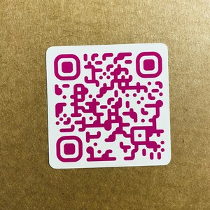 May include: A square white sticker with a magenta QR code. The QR code is composed of various magenta shapes and squares. The sticker has rounded corners and is set against a brown background.
