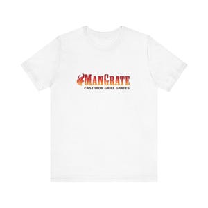 Puede incluir: Camiseta blanca con el texto "MANCRATE" en naranja y rojo, con un diseño de llama. Debajo, se lee "CAST IRON GRILL GRATES". La camiseta está hecha de un material suave.