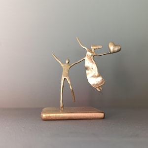 Puede incluir: Una escultura de metal color oro que representa dos figuras. Una figura está de pie con los brazos levantados, mientras que la otra parece estar flotando y sosteniendo un corazón. La escultura está montada sobre una base rectangular, creando una sensación de movimiento y emoción.