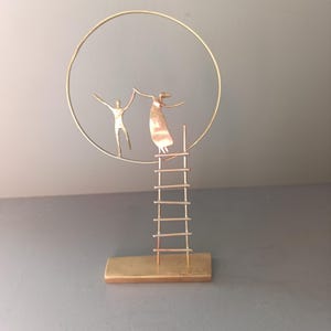 Puede incluir: Escultura de metal dorado con dos figuras estilizadas dentro de un marco circular. Una figura está sobre una escalera, extendiendo la mano hacia la otra. La escultura está montada sobre una base rectangular, creando una sensación de movimiento ascendente.