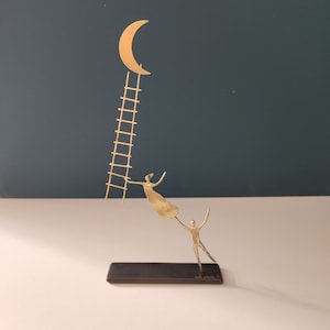 Op de afbeelding: Een gouden metalen sculptuur van twee figuren die naar een halve maan op een ladder reiken. Het beeld staat op een zwarte basis.