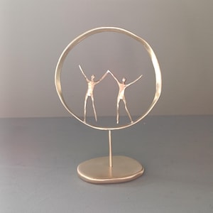Puede incluir: Una escultura de metal dorado con dos figuras estilizadas con los brazos levantados, unidas por las manos, dentro de un marco circular. La escultura está montada sobre una pequeña base ovalada, creando una obra de arte abstracta moderna.