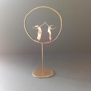 Puede incluir: Una escultura de metal dorado que presenta dos figuras estilizadas dentro de un marco circular. Las figuras parecen estar tomadas de la mano, con los brazos levantados. La escultura está montada sobre un soporte delgado y una base redonda.