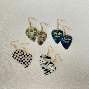 May include: A set of six guitar pick earrings. The picks are in various colors and patterns, including black and white checkered, green and white marbled, and blue and white marbled. Each pick has the word "Fender" printed on it, with the size of the pick indicated below the word. The earrings are made of gold-colored metal.