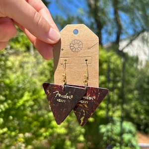May include: A pair of gold earrings featuring a brown and gold Fender guitar pick with the text "Fender Heavy" on each pick.