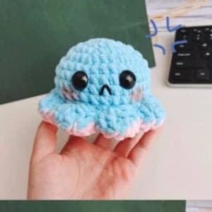 Könnte beinhalten: Zwei gehäkelte Octopus-Plüschtiere, eines blau und eines rosa, mit gestickten Augen und lächelnden Mündern. Die Plüschtiere werden in den Händen von zwei verschiedenen Personen gehalten.