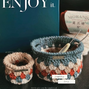 May include: Two handmade crochet baskets in shades of blue, beige, and red. The larger basket has a blue rim and contains crochet hooks. The text "ENJOY it" is at the top, and "Relax time" is at the bottom.