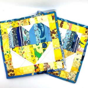 May include: Two yellow and blue patchwork fabric pot holders with a heart shape in the centre. The heart is made of blue and white fabric with a floral pattern.