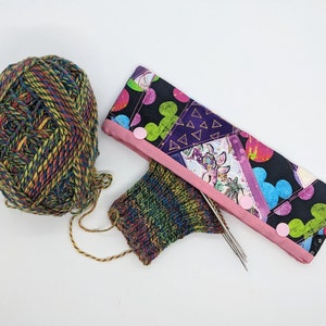 May include: A ball of multi-coloured yarn, a partially knitted sock, and two knitting needles are inside a colourful fabric needle case with pink trim and pink buttons.