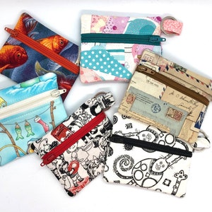 May include: A selection of small, square fabric coin purses with zippers and key rings. The purses display diverse patterns, including goldfish, patchwork, polka dots, vintage letters, and whimsical black and white cat designs. Each purse has a different coloured zip.