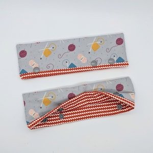 May include: Two gray fabric wristlets with red and white chevron patterns. The wristlets have a gray fabric lining with a yarn and needle design. The wristlets have three silver snaps.