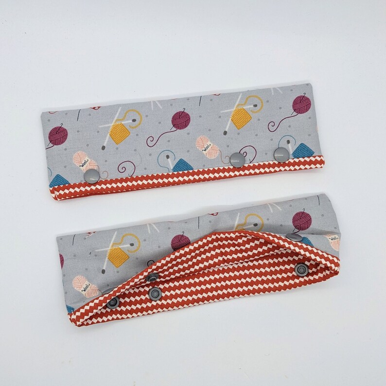 May include: Two gray fabric wristlets with red and white chevron patterns. The wristlets have a gray fabric lining with a yarn and needle design. The wristlets have three silver snaps.