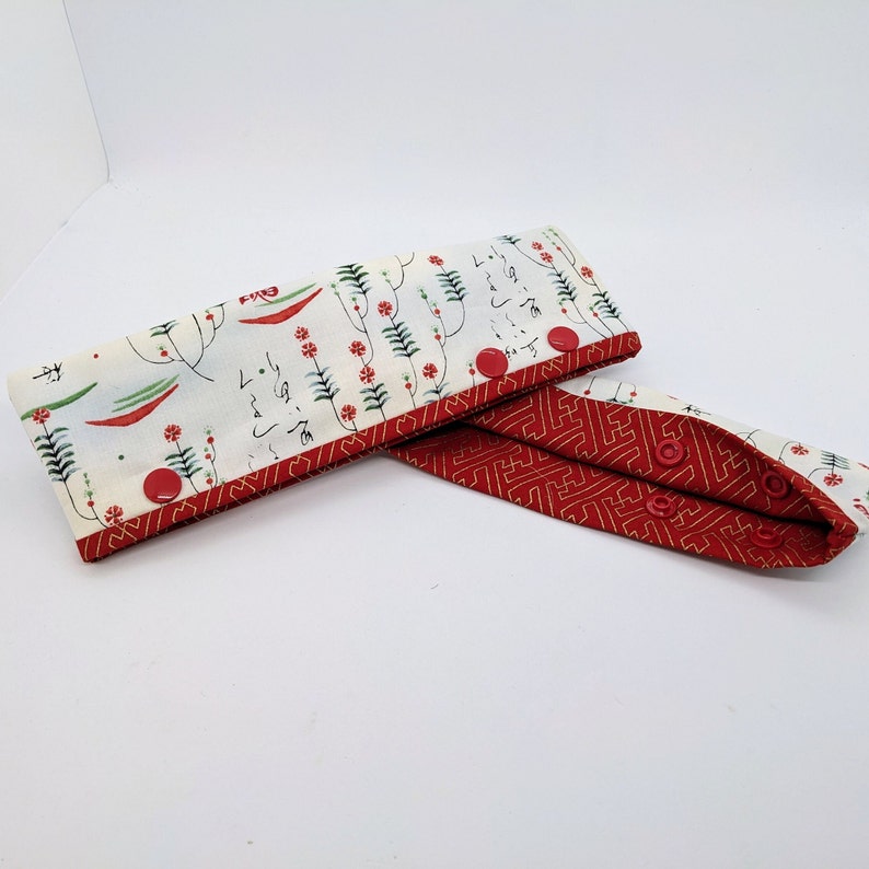 May include: A white fabric headband with red and green floral print and Japanese characters. The headband has a red fabric lining with a geometric pattern and two red buttons.