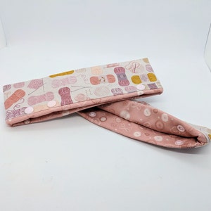 May include: A pink and white fabric headband with a pattern of yarn balls and knitting needles. The headband has two white buttons on each side.
