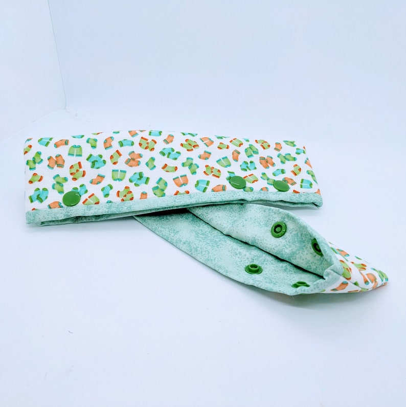 May include: A white fabric headband with a green and orange print of socks. The headband has green snap buttons and is folded in half.