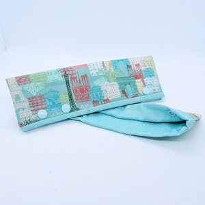 May include: A light blue fabric baby bib with a white and colorful print of Parisian landmarks. The bib has three white snap buttons on the side.