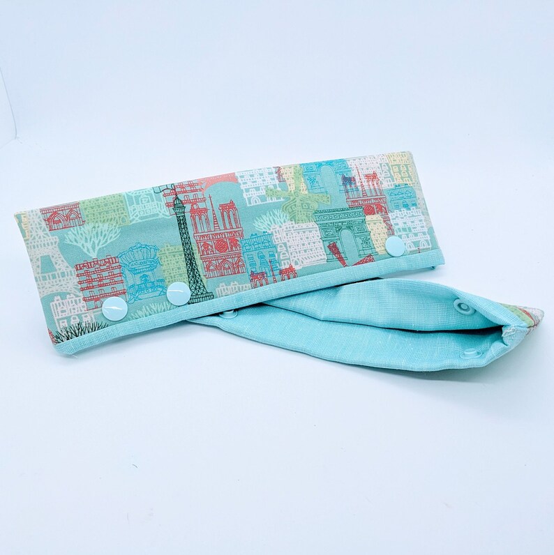 May include: A light blue fabric baby bib with a white and colorful print of Parisian landmarks. The bib has three white snap buttons on the side.