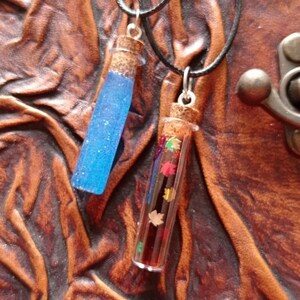 Könnte beinhalten: Zwei Halsketten mit Glasfläschchen und Korken. Eine Flasche enthält blaue Glitzerflüssigkeit, die andere farbige Konfetti in Blattform. Beide Fläschchen hängen an schwarzen Kordeln. Der Hintergrund ist eine strukturierte braune Oberfläche.