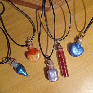 Peut inclure: Plusieurs colliers pendentifs avec de petites bouteilles décoratives et des bouchons en liège. Les bouteilles contiennent des substances colorées et scintillantes dans des tons de bleu, orange, rouge et violet. Chaque bouteille est suspendue à un cordon noir.