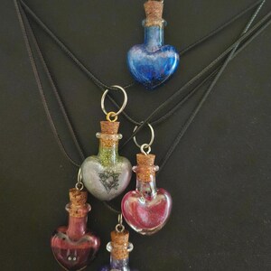 Può includere: Collane con ciondoli in vetro a forma di cuore, riempiti di liquidi colorati e tappi di sughero. I ciondoli sono blu, rossi, viola e argento, con un ciondolo d'argento su uno. Le collane sono su cordini neri.