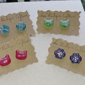 Op de afbeelding: Vier paar handgemaakte oorstekers op bruine kaarten. De oorbellen hebben de vorm van kattengezichten en pootafdrukken, in blauw, groen, roze en paars. De tekst "L'athétique Coffeuse" staat op elke kaart.