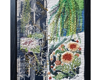 May include: A framed artwork featuring a patchwork of fabric with embroidered floral designs. The artwork includes a black and white fabric with a floral pattern, a white fabric with green embroidered leaves, and a white fabric with orange and green embroidered flowers. The artwork is signed "Lili" in the bottom right corner.